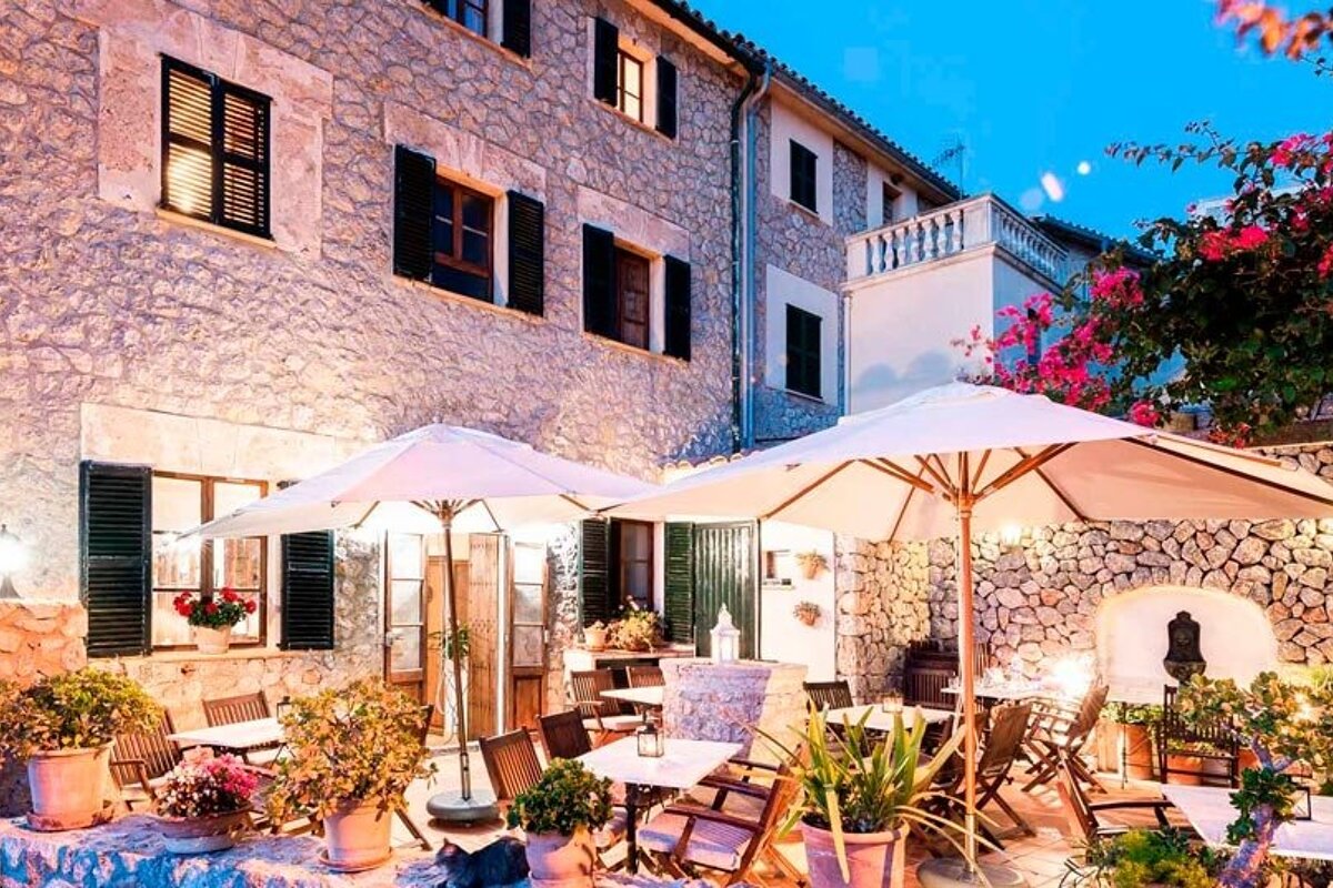 Cozy Mediterranean stone patio at dusk with dining tables, white umbrellas, potted plants, and vibrant pink bougainvillea under soft lights.