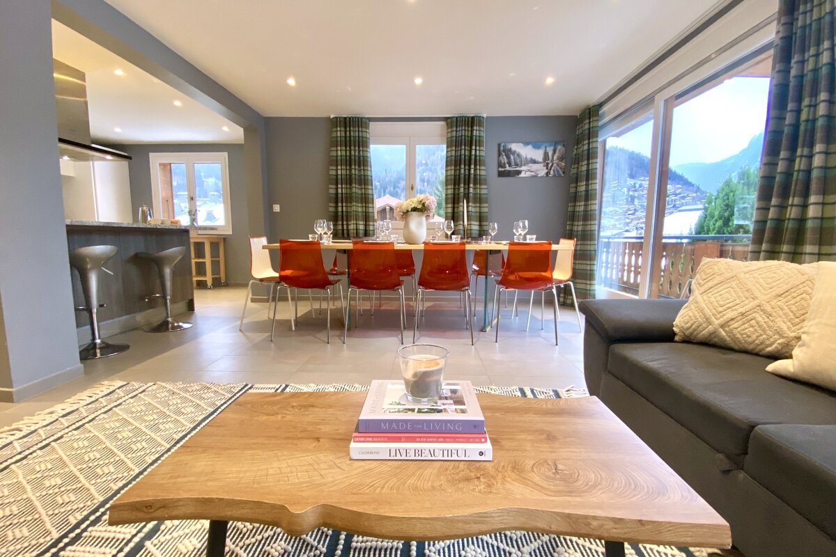 A stylish, modern open-plan living/dining room featuring a table with bright orange chairs, a cozy sofa, and a balcony with mountain views.