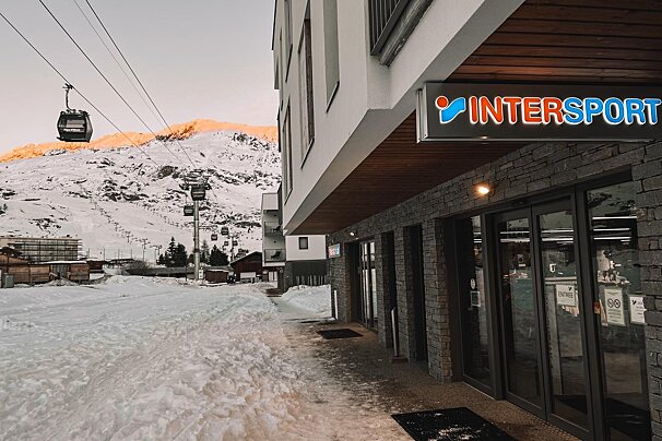 An Intersport store sits in a snowy ski resort, with gondolas and sun-kissed mountains in the background.