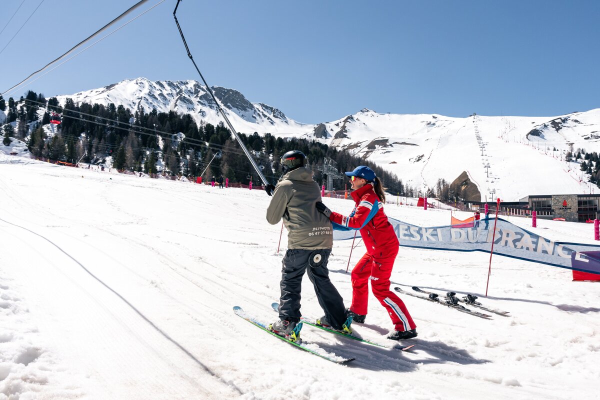 A ski instructor in red guides a student on a T-bar lift up a sunny, snowy mountain slope, with alpine trees and peaks in the background.