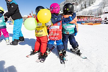 Three young children in ski gear, helmets, and Club Piou Piou vests stand on a snowy slope, holding colorful balloons during a ski lesson.