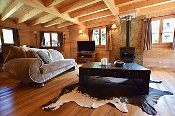 A cozy wooden chalet living room featuring a curved chaise, dark coffee table on a cowhide rug, and a rustic wood stove. Warm and inviting.