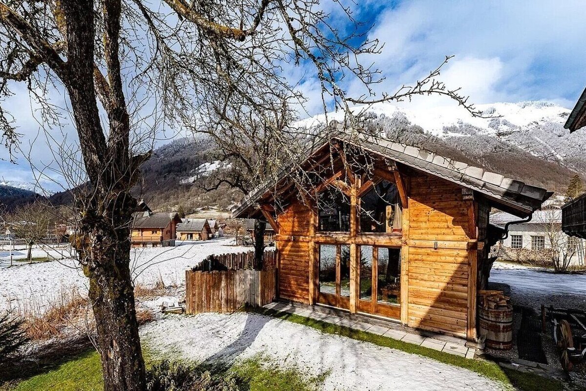 A beautiful wooden chalet sits in a snowy landscape with bare trees and majestic snow-capped mountains under a blue sky.