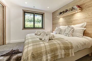 Cozy bedroom with a wooden feature wall, large bed with plaid throw and robes. A window overlooks greenery, with an animal hide rug.