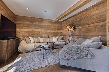 A cozy chalet living room with warm wood-paneled walls, plush grey sectional sofas, a shaggy rug, and an industrial floor lamp.
