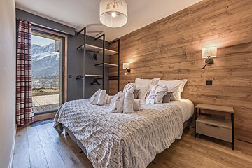 Cozy, modern bedroom with wood walls and a balcony offering stunning mountain views. Robes and towels await on the neatly made bed.