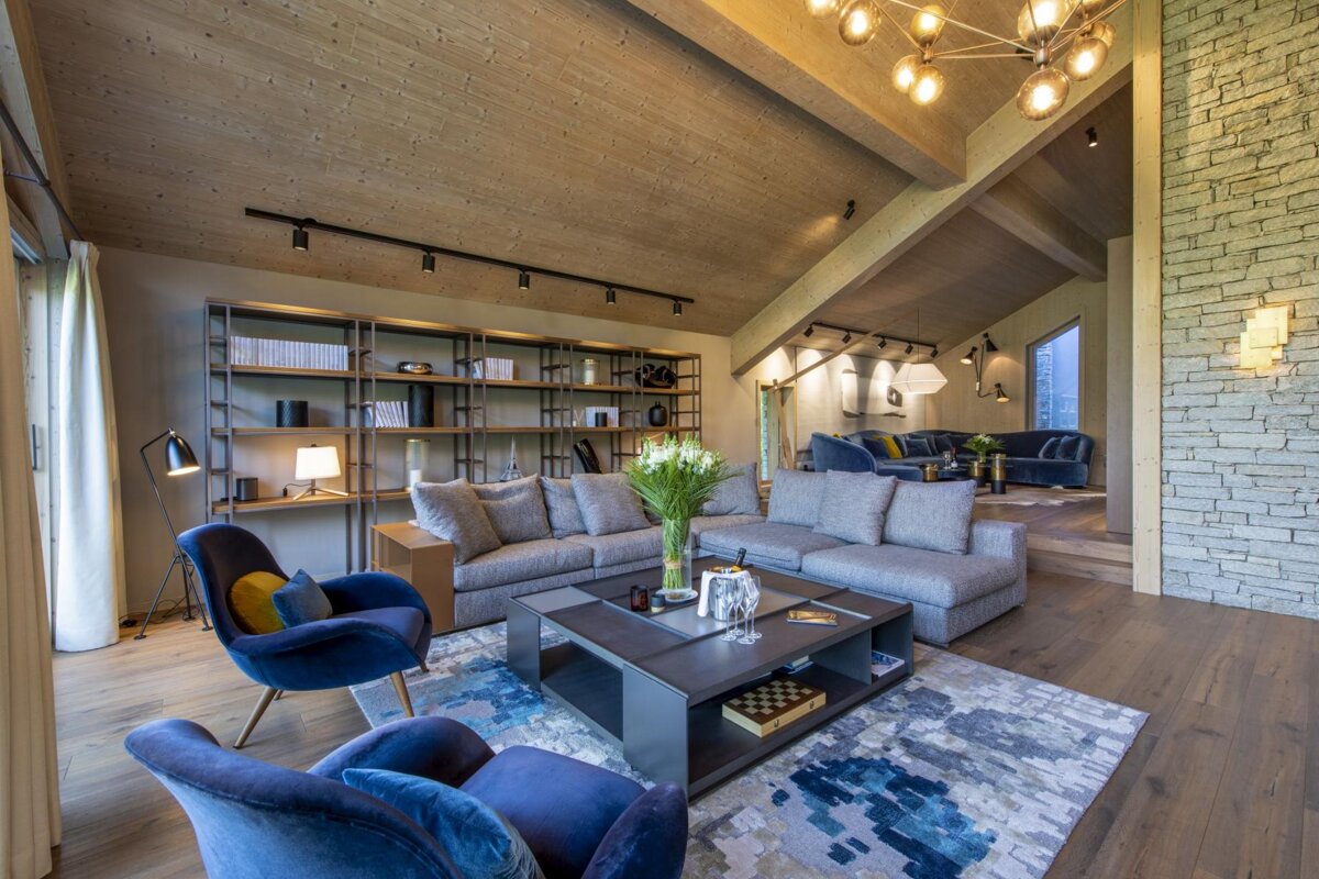 A stylish living room features a slanted wooden ceiling, grey sofas, blue accent chairs, a modern coffee table, a bookshelf, and chic lighting.