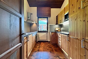 A narrow, rustic kitchen with natural wood cabinets, dark counters, hexagonal floor tiles, and a window overlooking greenery.