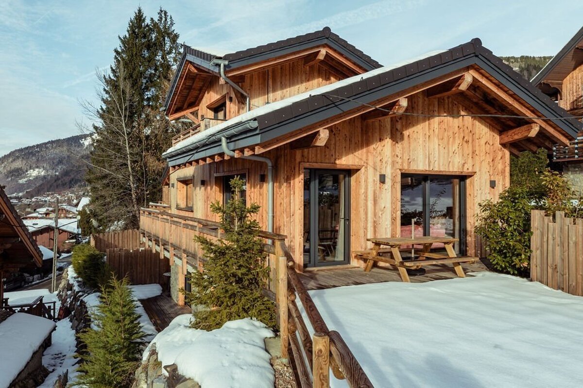 A rustic wooden chalet with a snowy deck, balcony, and dark roof, nestled in a mountain village during winter under a clear sky.