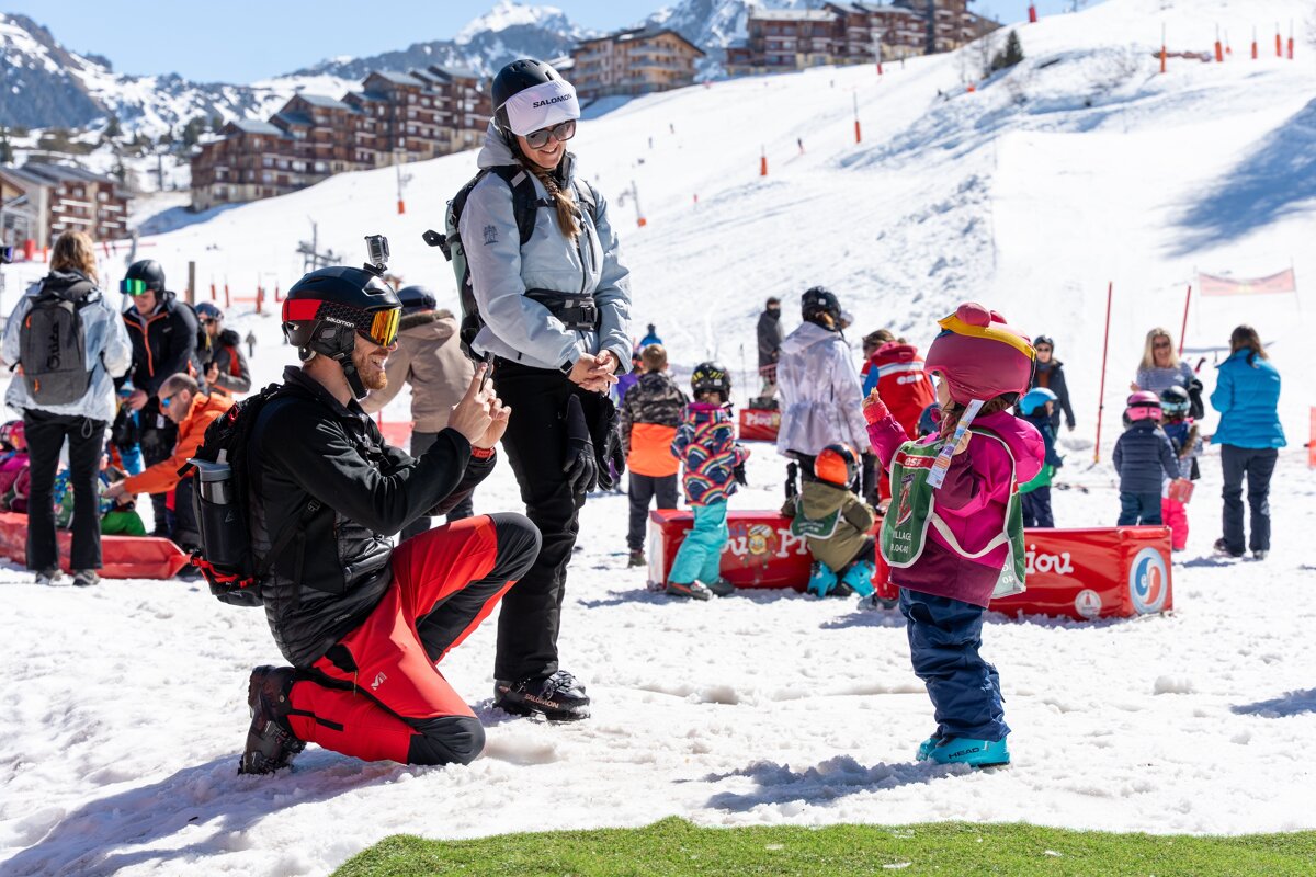 A man kneels to photograph a child on a snowy ski slope, with a woman smiling nearby. Other adults and children are in the background.