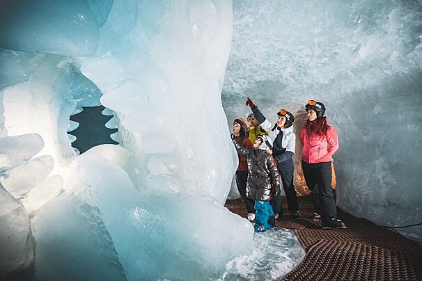A group of people, including a child, explore a breathtaking blue ice cave, looking up in awe at its magnificent frozen formations.