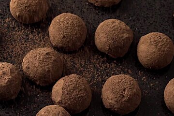 Close-up of brown, cocoa-dusted chocolate truffles on a dark surface. More cocoa powder falls onto them and is scattered around.