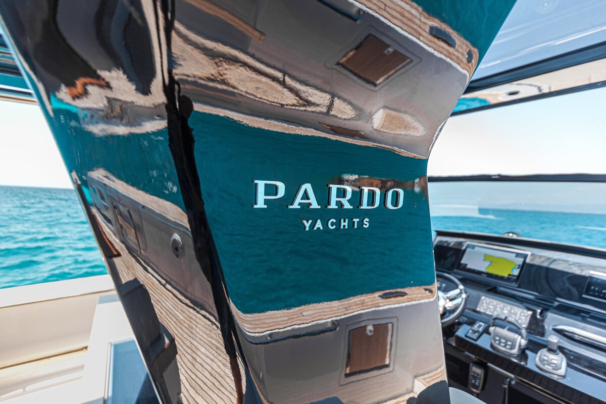 Close-up of the PARDO YACHTS logo on a shiny yacht panel, reflecting the sea and deck with the helm in the background.