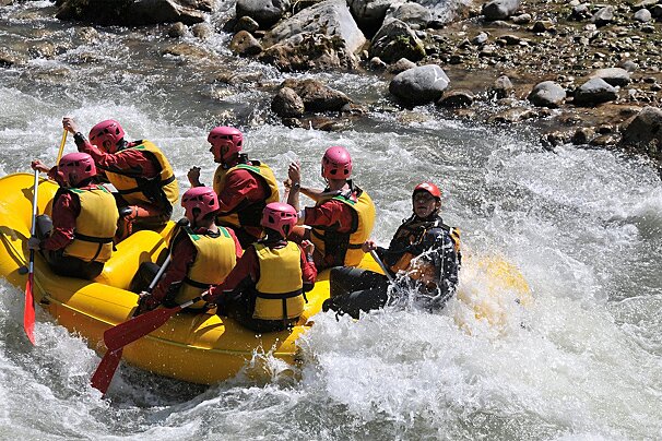 Seven people in a yellow raft, wearing helmets and life vests, are white water rafting through rapids on a rocky river.