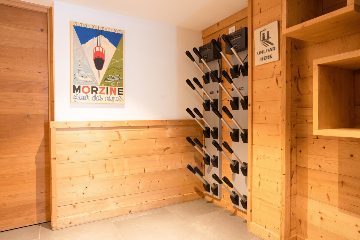 A cozy ski room with wooden paneling, a vintage Morzine poster, and a wall-mounted ski boot drying rack with an 
