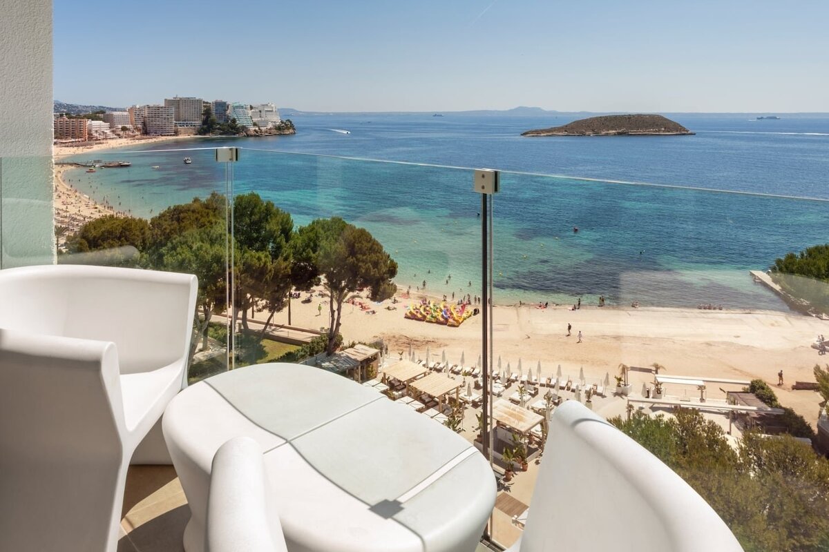 A bright balcony with white furniture overlooks a bustling sandy beach and clear turquoise sea, with distant coastal buildings and an island under a sunny sky.
