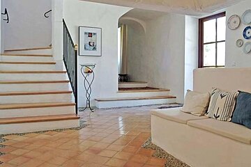 A bright, rustic interior with terracotta tiled floor, white walls, a staircase, and a cushioned built-in bench. Features an arched doorway and window.
