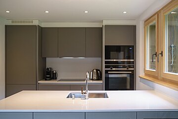 A modern kitchen with sleek gray cabinets, a white island with a sink, built-in appliances, and natural wood-framed windows.