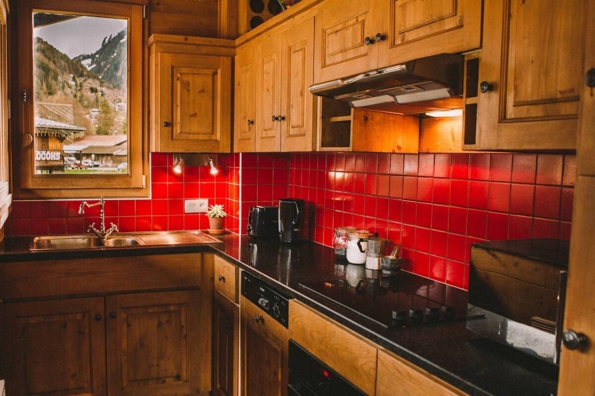 A rustic kitchen features warm wooden cabinets, a bright red tiled backsplash, black counters, and a window revealing a mountain village.