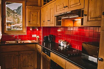 A rustic kitchen features warm wooden cabinets, a bright red tiled backsplash, black counters, and a window revealing a mountain village.