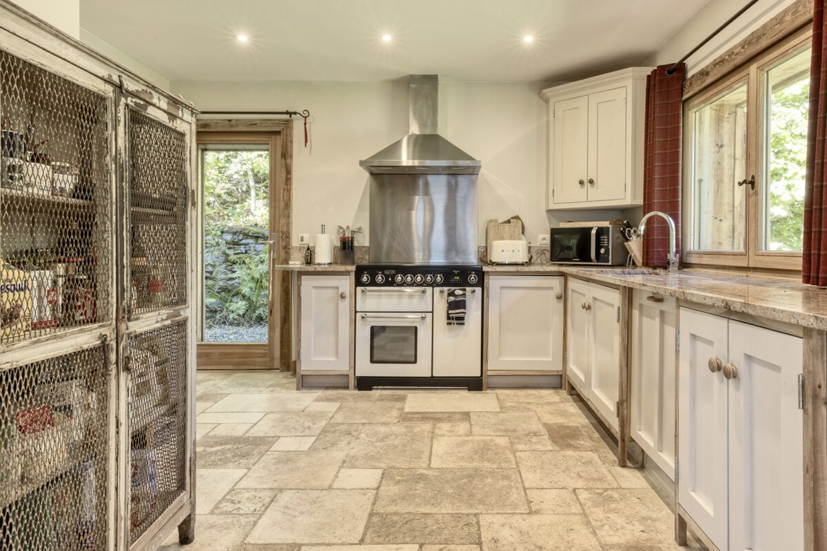A rustic kitchen with light stone floors, white cabinets, a large range cooker, and a distressed wire-mesh pantry. A door opens to an outdoor garden.