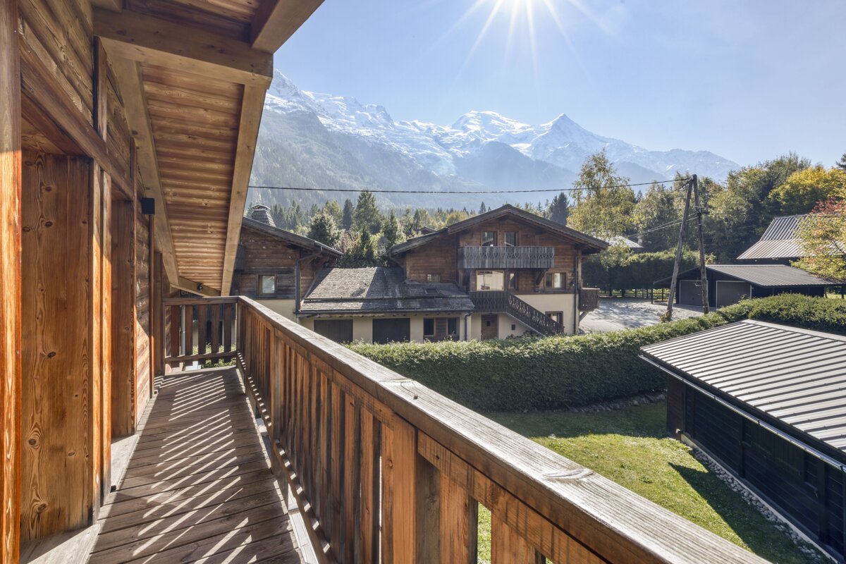 Sunny view from a wooden balcony revealing charming chalet homes, lush greenery, and majestic snow-capped mountains under a bright blue sky.