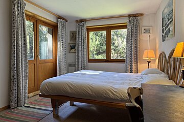 A sunlit bedroom features a bed with white linen, wooden doors and windows opening to a forest view, patterned curtains, and warm lamps.