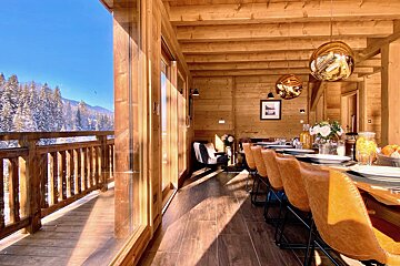 A warm, sunlit wooden dining room with a long table and a stunning view of snowy mountains and pine trees from a balcony.