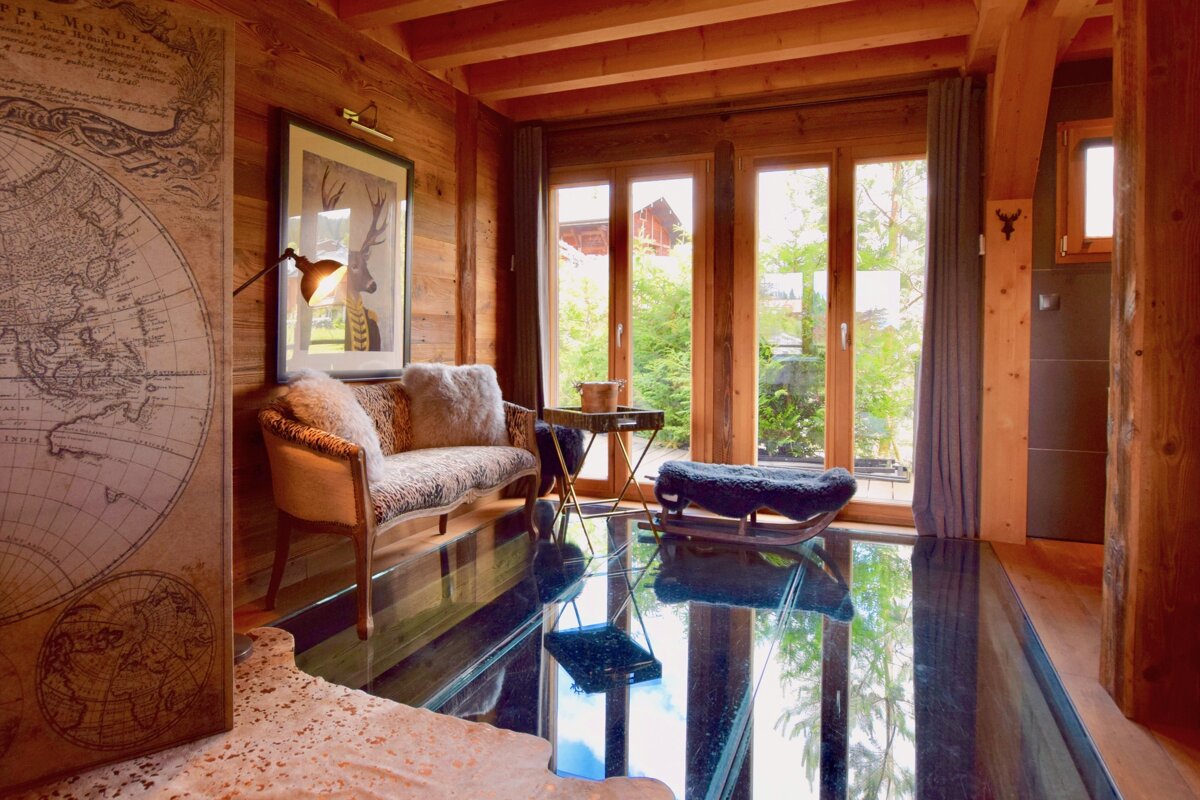 A cozy, rustic chalet room features warm wood paneling, large windows, a reflective floor, and a comfortable sofa with faux fur pillows.