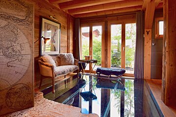 A cozy, rustic chalet room features warm wood paneling, large windows, a reflective floor, and a comfortable sofa with faux fur pillows.