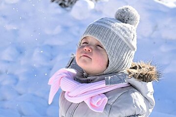 A bundled toddler in a grey pom-pom hat and pink scarf looks up at falling snowflakes in a sunny winter landscape.