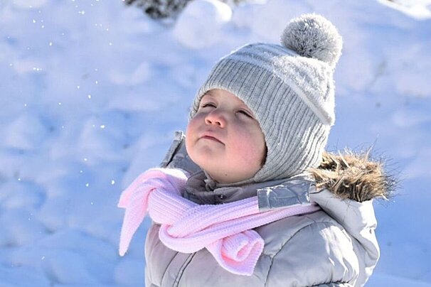 A bundled-up child in a grey hat and pink scarf looks up with closed eyes, enjoying the falling snow in a bright winter scene.