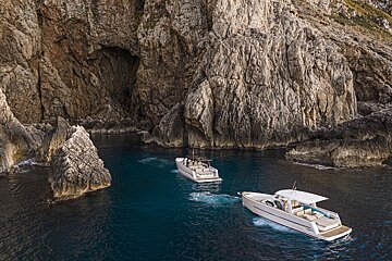 Two sleek modern boats cruise on dark blue water past dramatic rocky cliffs and a cave entrance.