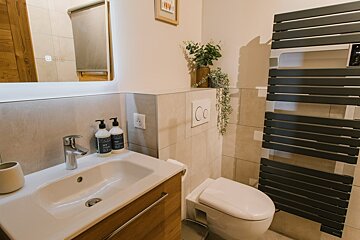 A modern bathroom with a white sink on a wood vanity, toilet, dark gray towel warmer, and decorative plants. Light tiles and walls complete the clean look.