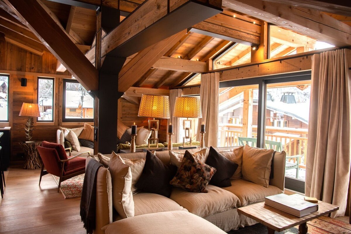 A warm, rustic wooden chalet living room with exposed beams, comfy sofas, and large windows looking out onto a snowy landscape.