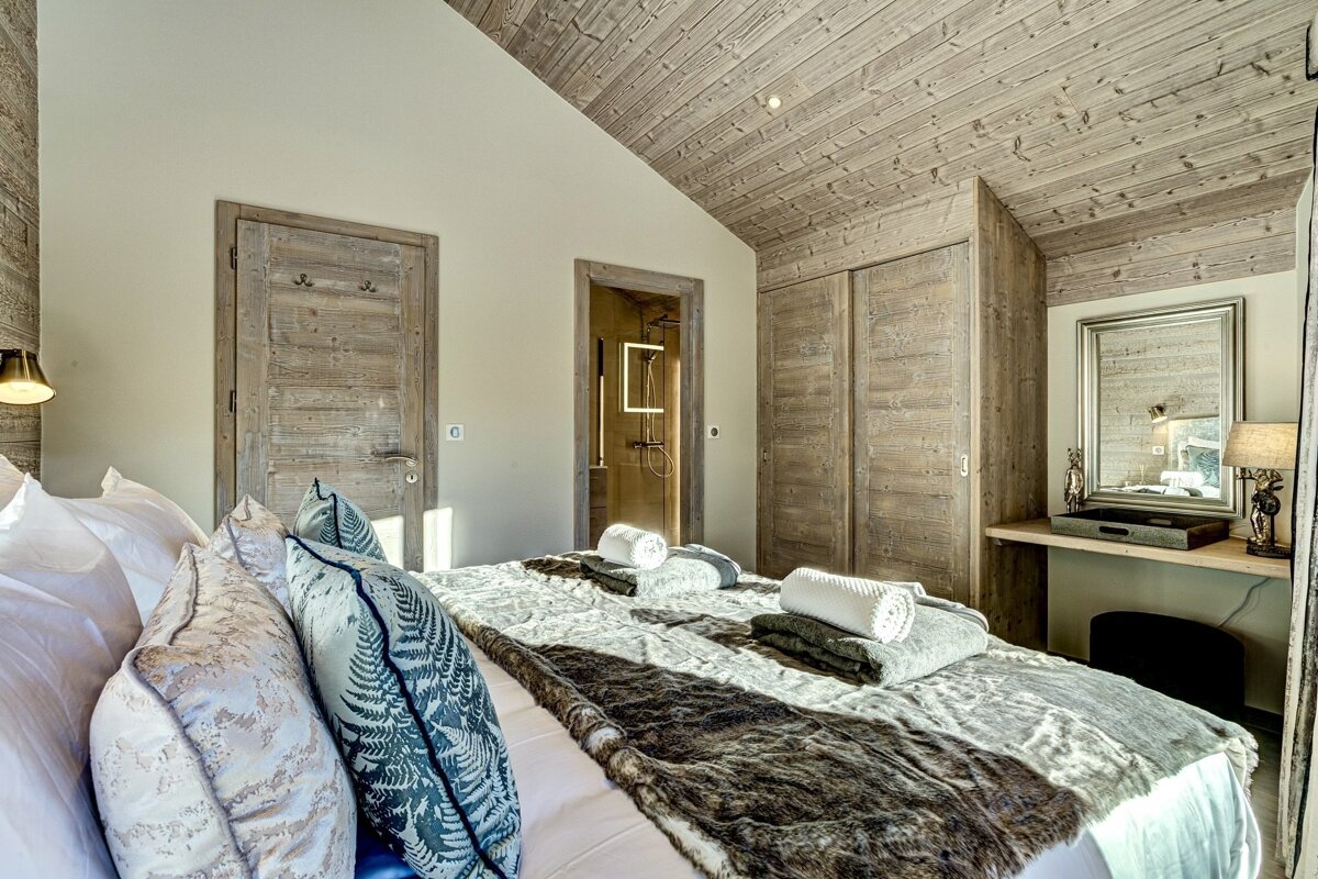 Cozy, rustic modern bedroom with a large bed, wood-paneled walls/ceiling, and a glimpse of an ensuite shower. Features a vanity with a mirror.