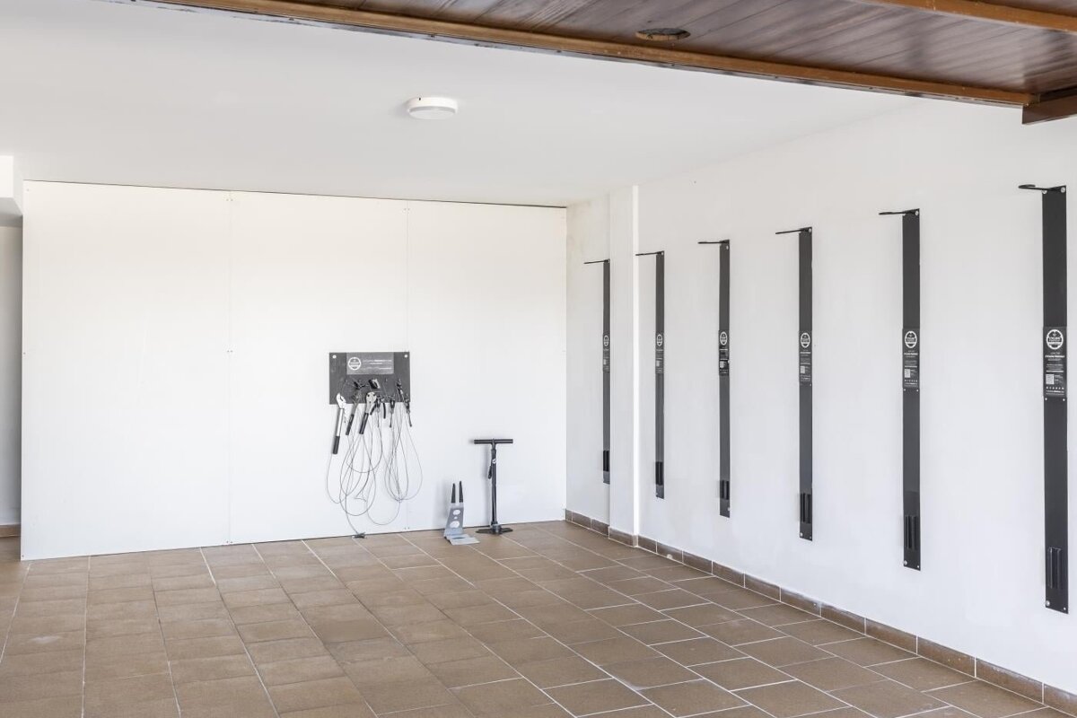 A bike repair station with tools, a floor pump, and several vertical bike racks against white walls and a tiled floor, under a wooden ceiling.
