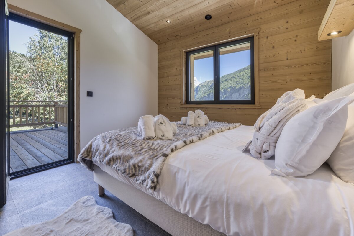 A cozy bedroom with a large bed, wooden walls/ceiling, and stunning views of mountains from a window, plus a balcony overlooking a forest.