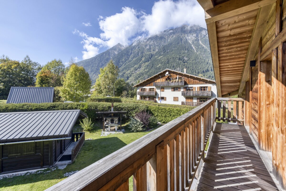 Sunny view from a wooden deck overlooking alpine chalets, lush greenery, and towering, cloud-kissed mountains on a bright day.