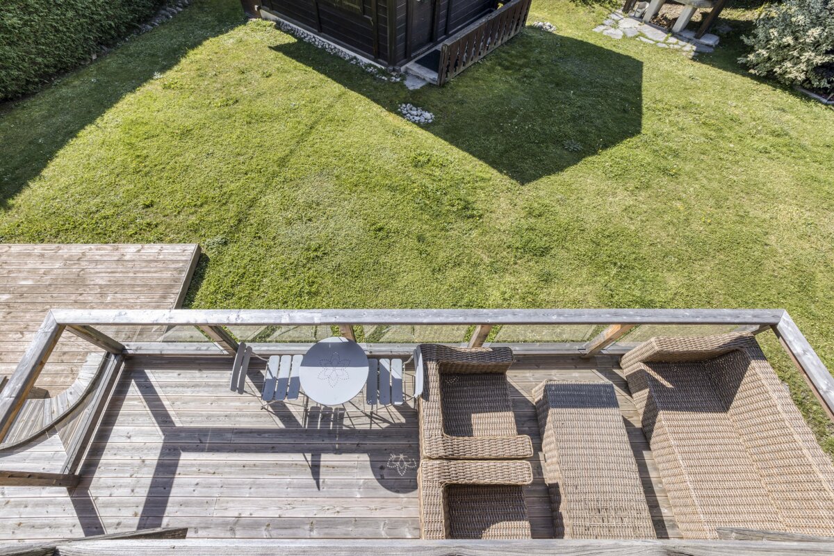 Overhead view of a sunny wooden deck with outdoor furniture (table, chairs, lounge beds) overlooking a green lawn and a small shed. Strong shadows.
