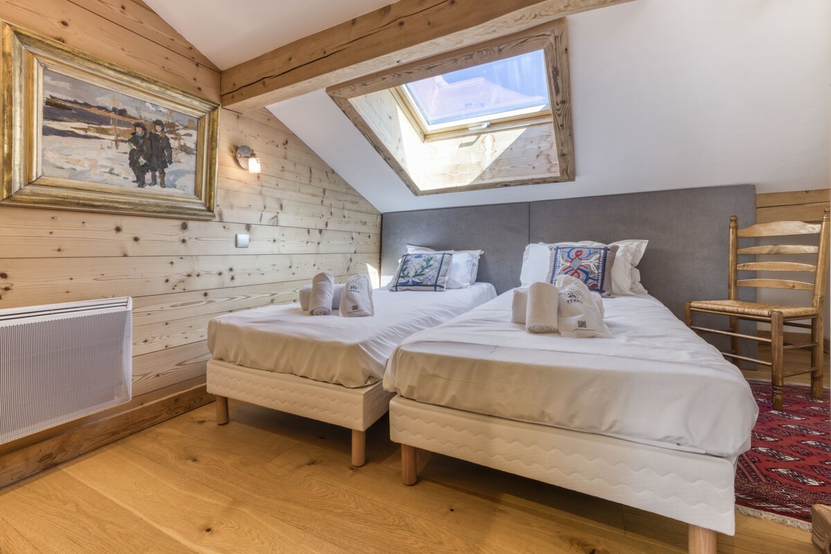A cozy attic bedroom with two twin beds, wood-paneled walls, a skylight, and a large painting, creating a rustic, inviting atmosphere.