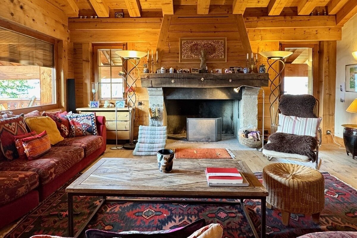 A cozy, rustic log cabin living room featuring a large stone fireplace, a red sofa with patterned pillows, and a wooden coffee table on a patterned rug.