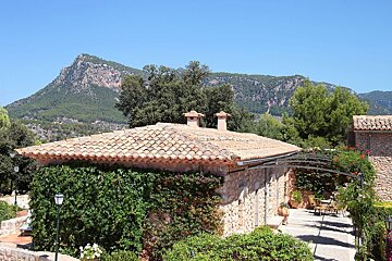 A rustic stone building with a red-tiled roof and ivy-covered walls, set against a backdrop of forested mountains under a clear blue sky.
