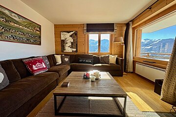 Cozy chalet living room with a brown sectional sofa, wooden coffee table, and stunning snowy mountain views from large windows. Sunny rustic interior.