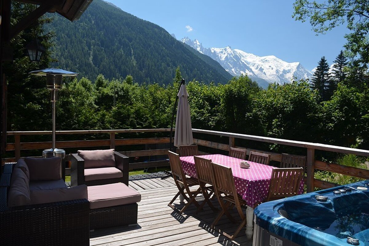 A sunny wooden deck with outdoor furniture, a hot tub, and a stunning view of snow-capped mountains and green forests.
