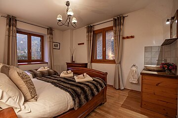 A cozy twin bedroom with wooden beds, faux fur throws, and clean towels. Features wooden windows, a charming chandelier, and a stone sink with flowers.