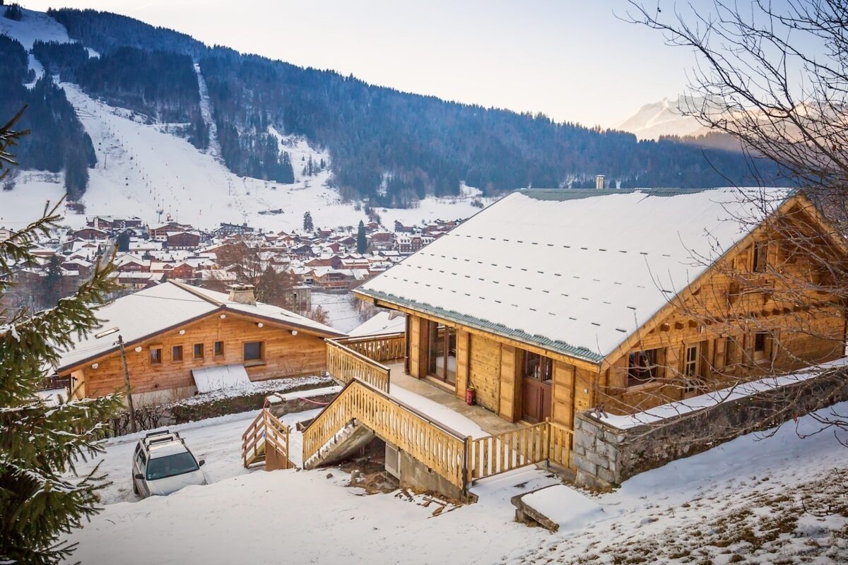 Snow-covered wooden chalets nestled in a mountain village, with ski slopes winding down a snow-laden peak in the background on a clear day.