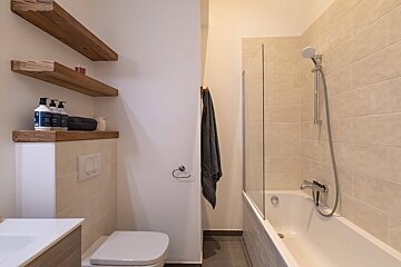 A modern, neutral-toned bathroom features a toilet, bathtub with a glass shower screen, and wooden floating shelves holding toiletries.
