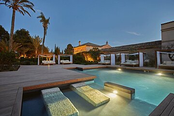 Dusk view of a luxurious resort pool with cabanas, palm trees, and illuminated historic buildings. Includes unique in-pool lounge chairs.