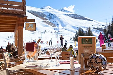 An outdoor dining table set with wine and glasses, overlooking sunny, snow-covered mountains with skiers at a cozy ski resort.